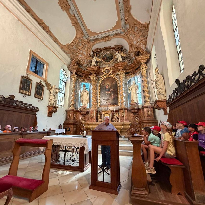 In einer Kirche steht ein Mann mit einem Mikrofon an einem Podest. Kinder sitzen auf Bänken vor ihm. Die Kirche ist mit aufwendigen Schnitzereien und Gemälden geschmückt.