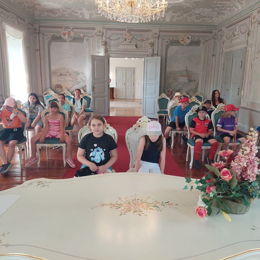 Eine Gruppe von Kindern sitzt in Stühlen um einen Tisch in einem kunstvollen Raum mit Blumentapeten und einem Kronleuchter.