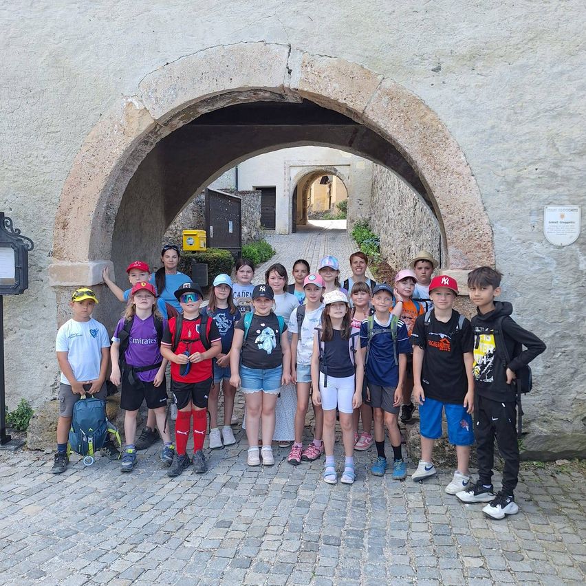 Eine Gruppe von Kindern und Erwachsenen steht auf Kopfsteinpflaster vor einem steinernen Bogen. Sie tragen Freizeitkleidung, Hüte und tragen Rucksäcke. Einige tragen Sonnenbrillen. Ein Schild ist an der Wand montiert.