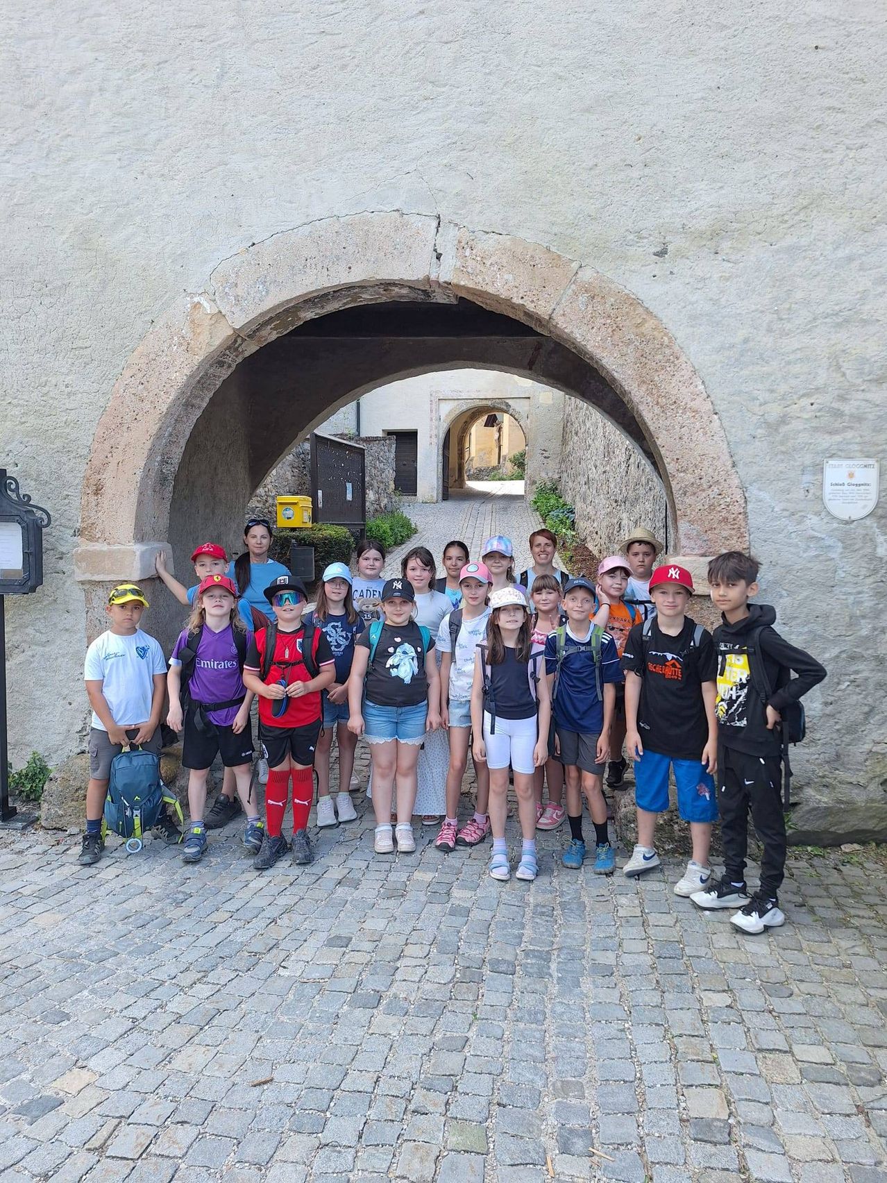 Eine Gruppe von Kindern und Erwachsenen steht auf Kopfsteinpflaster vor einem steinernen Bogen. Sie tragen Freizeitkleidung, Hüte und tragen Rucksäcke. Einige tragen Sonnenbrillen. Ein Schild ist an der Wand montiert.