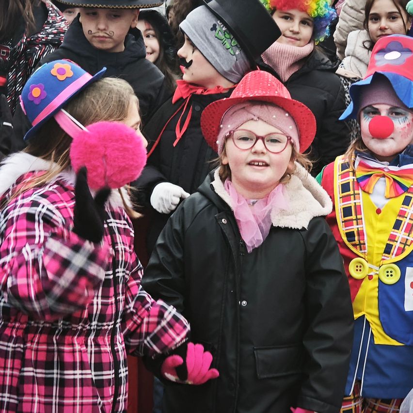 Eine Gruppe von Kindern in Kostümen, einschließlich Hüten und Handschuhen, steht draußen zusammen. Eines trägt einen roten Hut, ein anderes hat einen rosa Handschuh, und ein Clown mit gemalten Nase ist ebenfalls anwesend.