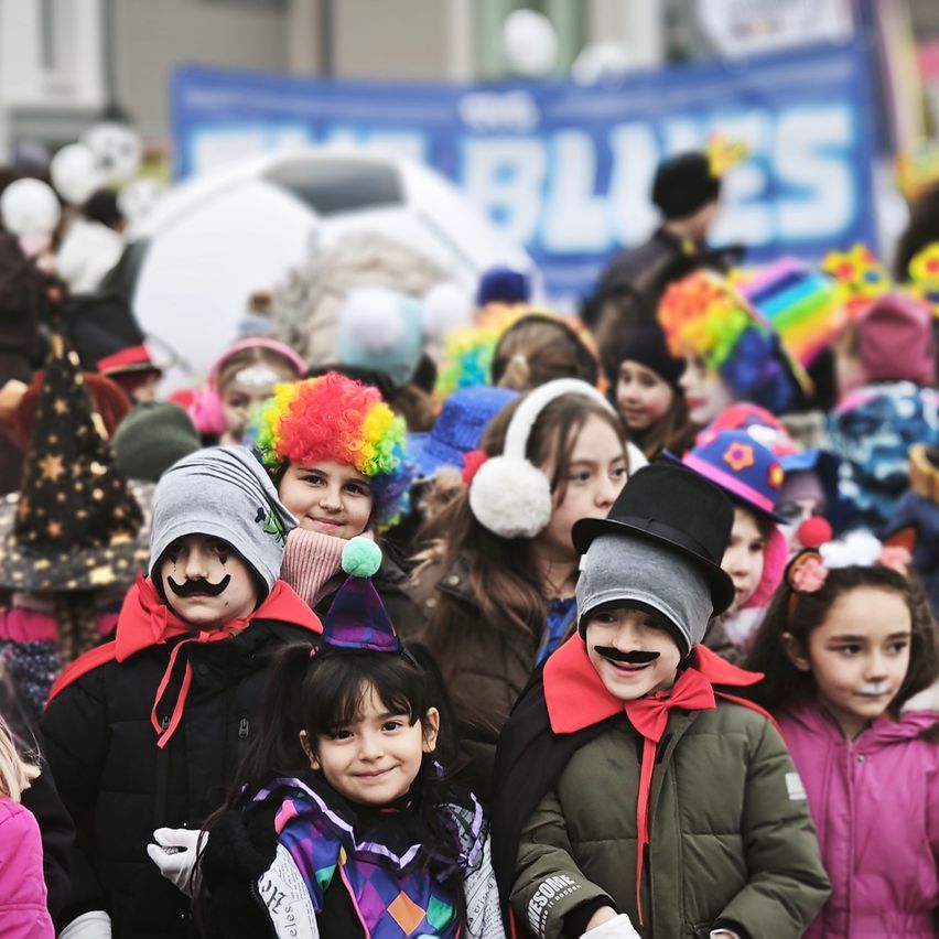 Eine Gruppe von Kindern in Kostümen, darunter ein Mädchen mit Schnurrbart, posiert für ein Foto auf einem Festival. Sie tragen verschiedene farbenfrohe Hüte und Jacken, einige haben Schminke im Gesicht. Im Hintergrund steht ein Banner mit der Aufschrift 'Blues'.