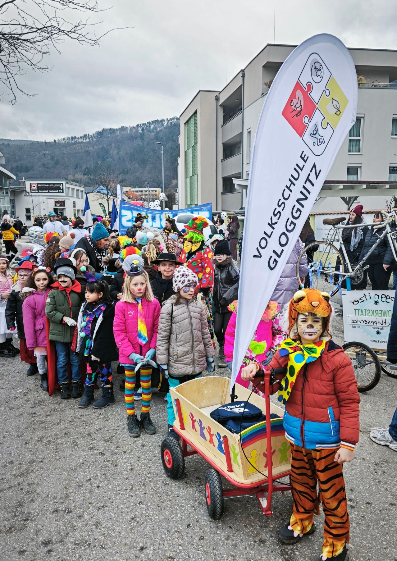 Eine Gruppe von Kindern in bunten Kostümen und mit Gesichtsbemalung nehmen an einem Umzug teil, mit einem Banner, das Volksschule Gloggnitzer liest. Vor ihnen sind Fahrräder geparkt, und Menschen in Winterkleidung stehen daneben und beobachten den Umzug.