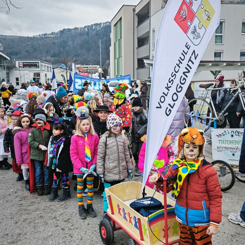 Eine Gruppe von Kindern in bunten Kostümen und mit Gesichtsbemalung nehmen an einem Umzug teil, mit einem Banner, das Volksschule Gloggnitzer liest. Vor ihnen sind Fahrräder geparkt, und Menschen in Winterkleidung stehen daneben und beobachten den Umzug.