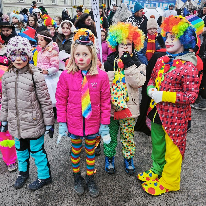 Mehrere Kinder in verschiedenen Kostümen, darunter Clowns, stehen auf einer Stadtstraße, wobei einige Gesichtsbemalung tragen und andere farbenfrohe Perücken und Schals haben.