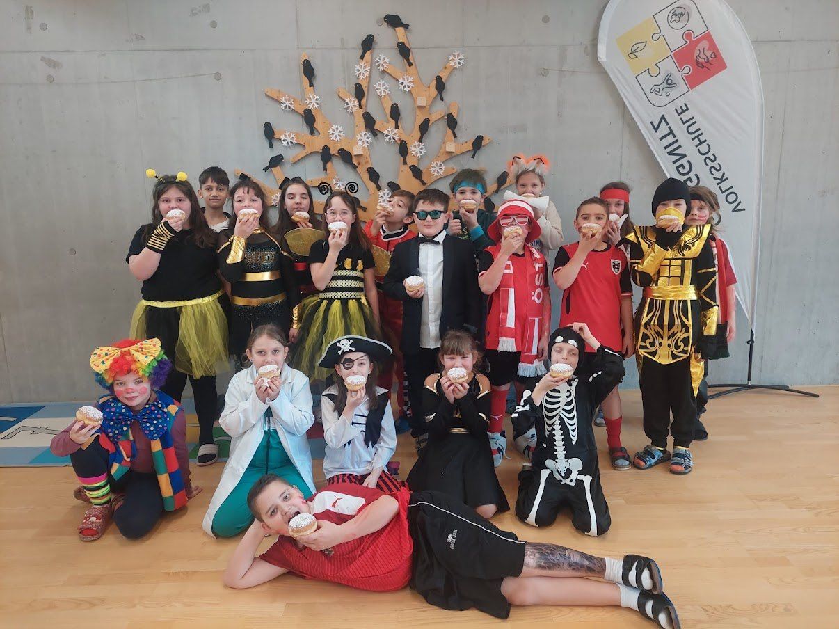 Eine Gruppe von Kindern in Kostümen posiert für ein Foto, alle halten Donuts und haben fröhliche Gesichter. Ein dekorativer Baum mit Schneeflocken und Vögeln steht hinter ihnen.