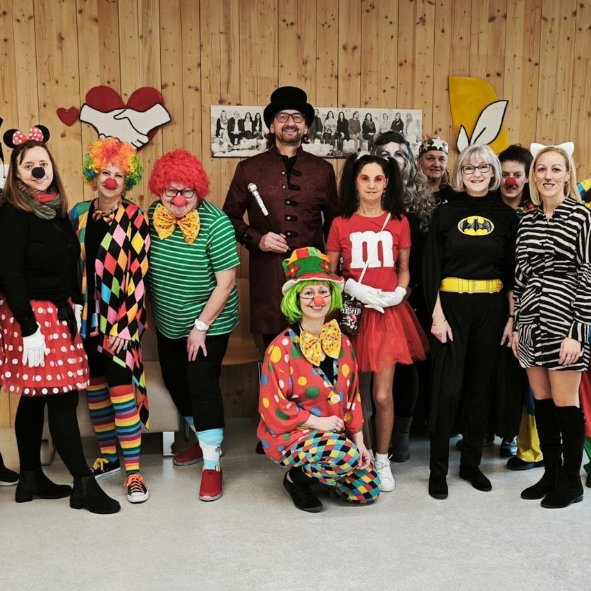 Eine Gruppe Erwachsener in bunten Kostümen posiert für ein Foto in einem Raum mit Holzwänden. Ein Mann in einem Clownskostüm hockt auf dem Boden.