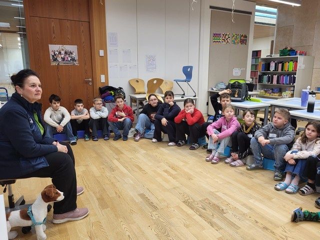 Eine Frau mit einem Hund sitzt auf einem Stuhl in einem Klassenzimmer, umgeben von Kindern, die im Kreis sitzen. Die Kinder lauschen aufmerksam. Hinter ihnen befinden sich Tische, Stühle und ein Regal mit bunten Gegenständen.