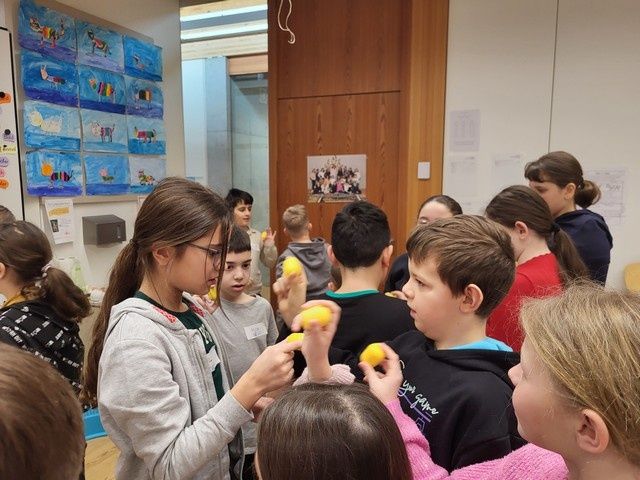 Eine Gruppe von Kindern hält gelbe Bälle und scheint ein Spiel zu spielen. Ein Mädchen mit Brille hält einen gelben Ball und scheint zu unterrichten.