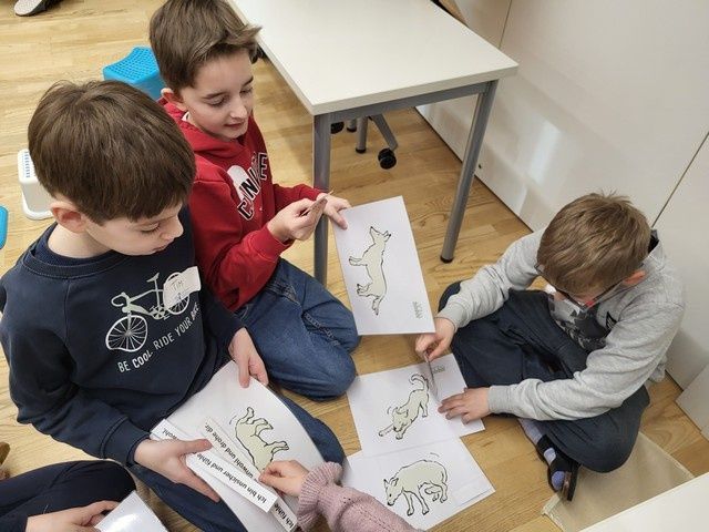 Vier Kinder sitzen auf einem Holzboden und sind mit einer Malaktivität beschäftigt. Jedes Kind hält ein Stück Papier mit einer Hundezeichnung in der Hand. Die Kinder tragen Freizeitkleidung. Im Hintergrund steht ein weißer Tisch.
