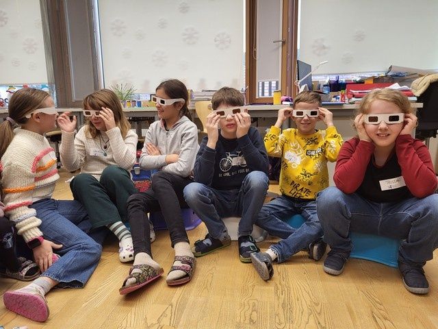 Eine Gruppe von Kindern sitzt auf blauen Hockern und trägt weiße Brillen mit schwarzen Rahmen. Sie lächeln und schauen etwas vor sich an. Sie tragen Freizeitkleidung und Turnschuhe. Der Raum hat einen Holzboden und eine weiße Wand mit Schneeflocken. Es gibt einen Schreibtisch mit einer Topfpflanze obenauf.