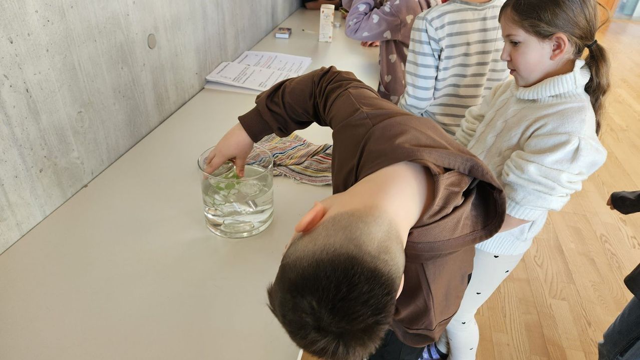 Ein Junge beugt sich über einen Tisch und beobachtet ein Glas mit Eiswürfeln. Hinter ihm sitzen zwei Kinder auf Stühlen. Papiere und eine Schachtel liegen auf dem Tisch.
