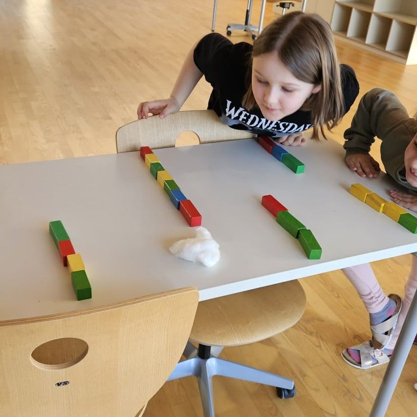 Zwei Mädchen spielen mit bunten Bauklötzen auf einem weißen Tisch in einem Klassenzimmer. Das Mädchen in Schwarz konzentriert sich auf die Blöcke, während das andere Mädchen lächelt.
