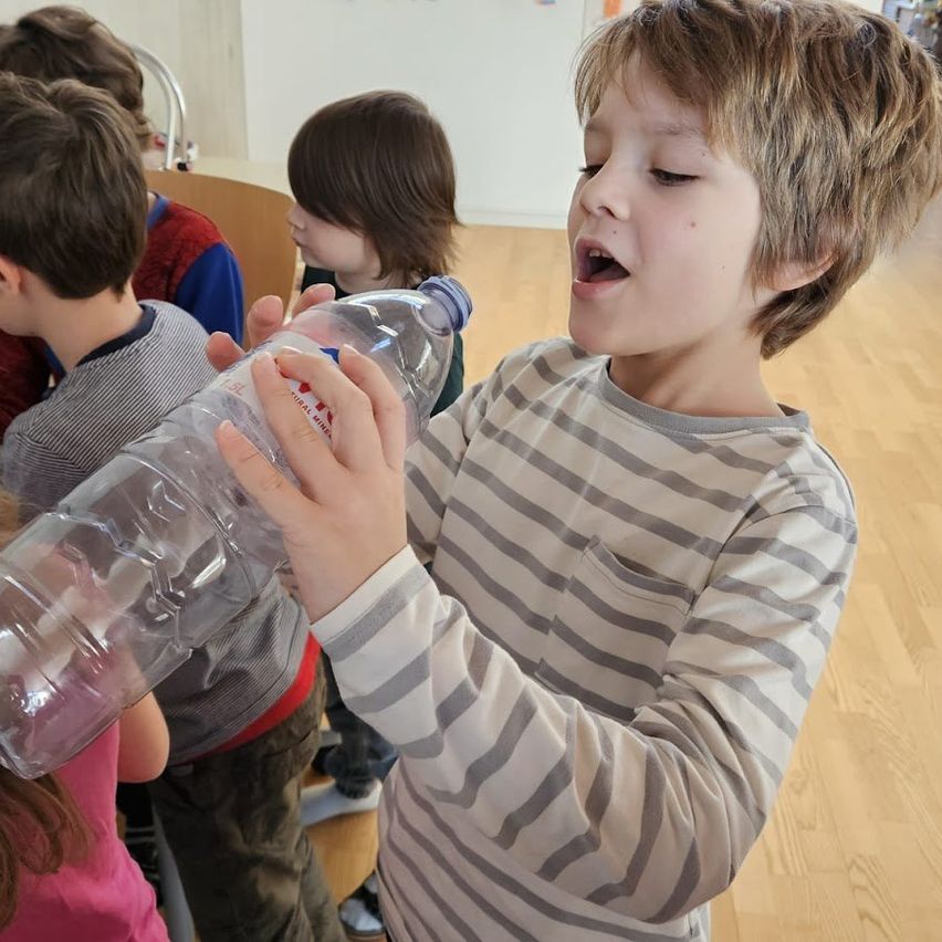 Ein junger Junge bläst in eine klare Plastikflasche im Klassenzimmer. Andere Kinder stehen hinter ihm.