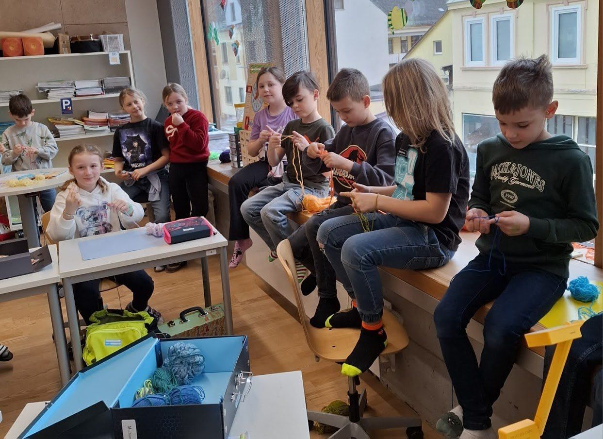 Eine Gruppe von Kindern, sowohl Jungen als auch Mädchen, sitzen um einen Tisch und auf einer Bank herum und stricken. Eine offene Kiste mit Garn steht auf dem Tisch vor ihnen, und sie halten alle Garn in den Händen.