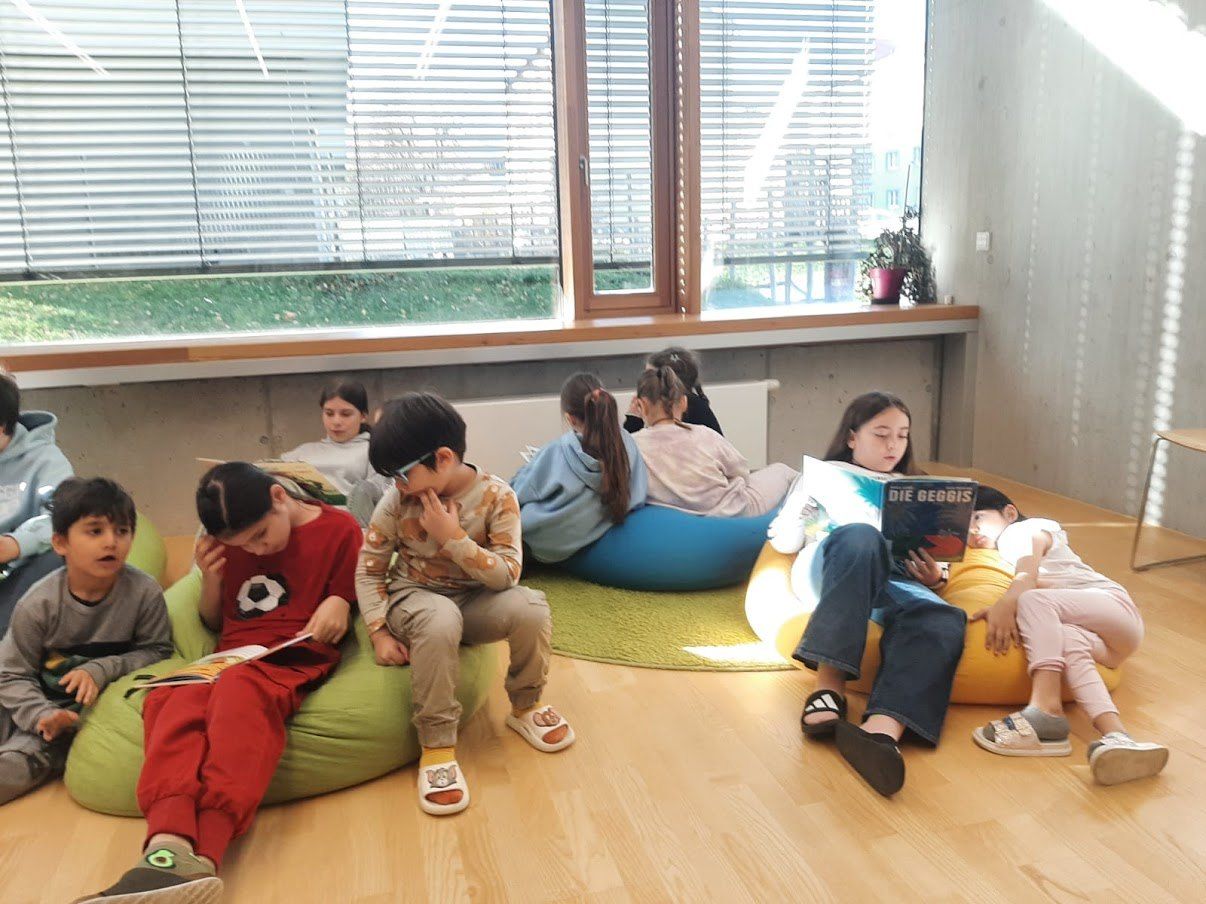 Eine Gruppe von Kindern sitzt auf Beanbags und liest Bücher. Sie sind auf einem Holzboden in der Nähe eines Fensters mit Jalousien.