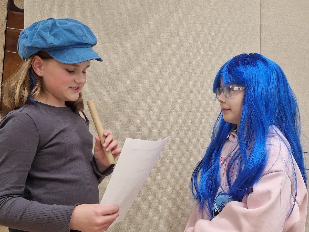 Zwei Mädchen stehen vor einer beigefarbenen Wand. Ein Mädchen hält ein Papier und einen Stock, das andere trägt eine blaue Perücke und eine Brille.