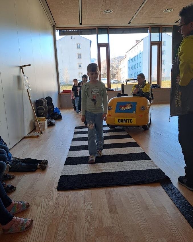 Ein kleines Mädchen läuft auf einem schwarz-weiß gestreiften Teppich in einem Raum mit Holzböden, einige Personen stehen herum und ein kleines gelbes Fahrzeug im Hintergrund.