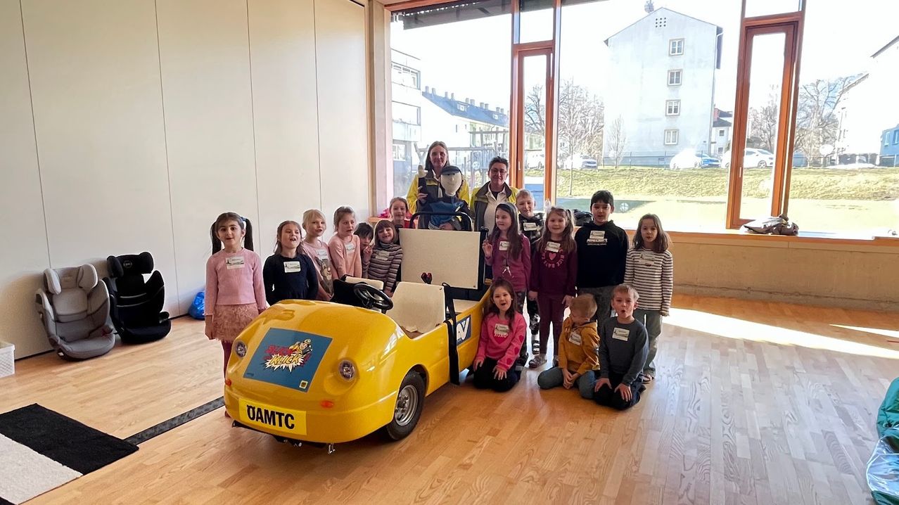 Eine Gruppe von Kindern und zwei Erwachsenen posiert für ein Foto mit einem gelben Spielzeugauto in einem Raum mit Holzböden. Das Auto hat 'OAMTC' auf der Rückseite. Die Kinder tragen Namensschilder.