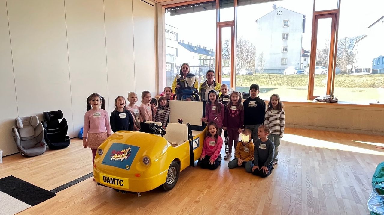 Eine Gruppe von Kindern und zwei Erwachsenen posiert mit einem gelben Spielzeugauto in einem Raum mit Holzboden und großen Fenstern, die einen Rasenbereich und Gebäude draußen zeigen.