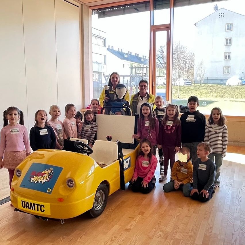 Eine Gruppe von Kindern und zwei Erwachsenen posiert mit einem gelben Spielzeugauto in einem Raum mit Holzboden und großen Fenstern, die einen Rasenbereich und Gebäude draußen zeigen.