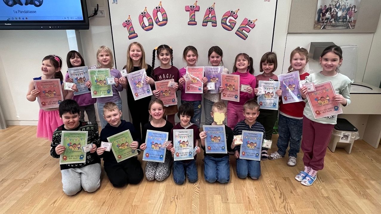 Mehrere junge Kinder posieren für ein Foto mit Urkunden vor einer Wand, die '100 Tage' steht. Sie alle haben ein Lächeln im Gesicht.
