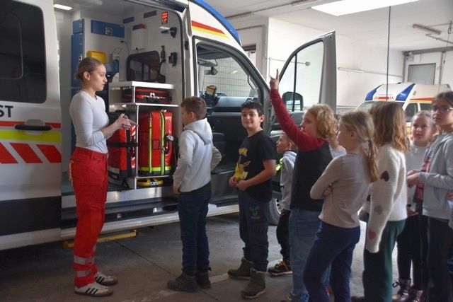Eine Frau in roter Uniform erklärt einer Gruppe von Kindern in einer Garage etwas über einen Krankenwagen.