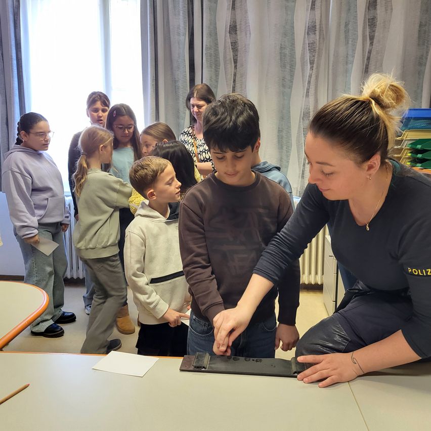 Eine Gruppe von Kindern und einer Frau in Polizeiuniform versammelt sich um einen Tisch. Die Frau demonstriert den Kindern etwas am Tisch, die neugierig zusehen.