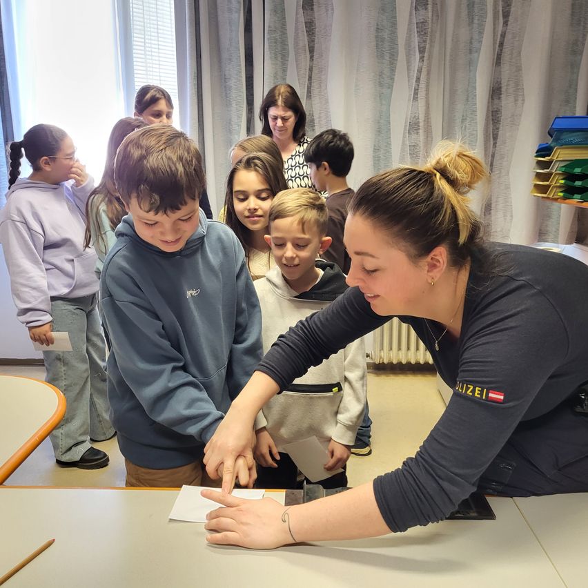 Eine erwachsene Frau in einem schwarzen Shirt unterrichtet junge Kinder, die um einen Tisch herum sitzen, einer hält ein Stück Papier.