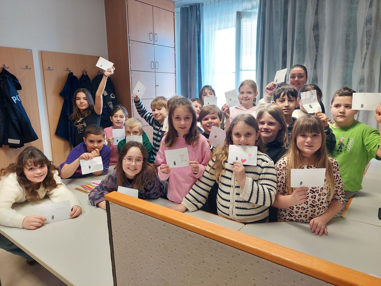 Ein Klassenzimmer voller Kinder, die kleine Papiere hochhalten, einige mit Zeichnungen, in einem Raum mit weißen Wänden, Holzschränken und blauen Vorhängen.