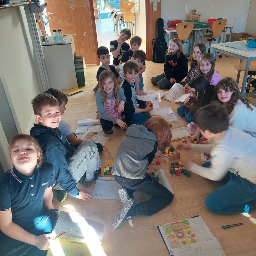 Eine Gruppe von Kindern sitzt auf dem Boden in einem Klassenzimmer und ist in einer praktischen Aktivität mit Bauklötzen engagiert. Einige schauen in Bücher, während andere spielen.