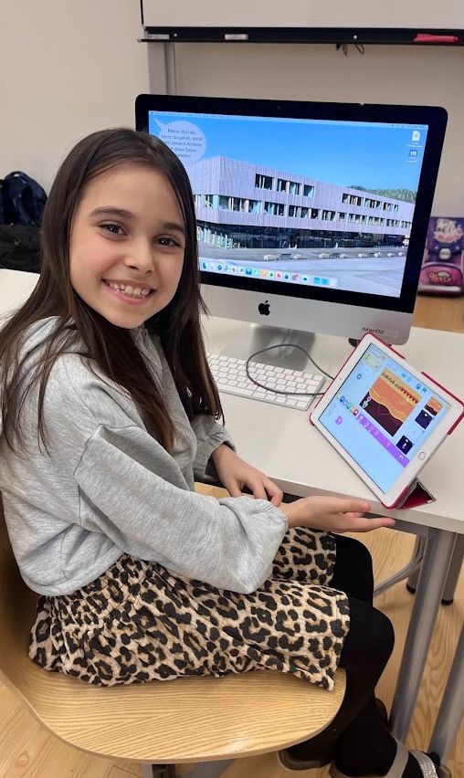 Ein Mädchen sitzt an einem Schreibtisch, lächelt in die Kamera, mit einem Computermonitor und einem Tablet vor sich.