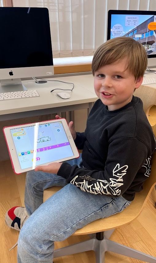 Ein junger Junge in einem schwarzen Sweatshirt und Jeans sitzt auf einem Stuhl und hält ein iPad mit einer Zeichen-App in der Hand.