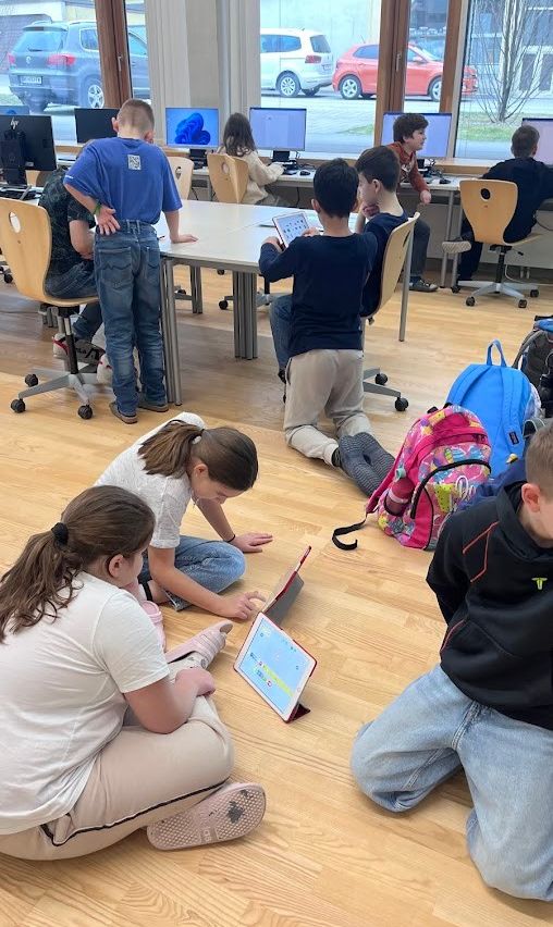 Schüler in einem Klassenzimmer verwenden Tablets und Laptops. Einige sitzen auf dem Boden, andere an Schreibtischen mit Monitoren. Rucksäcke und Schuhe sind verstreut.
