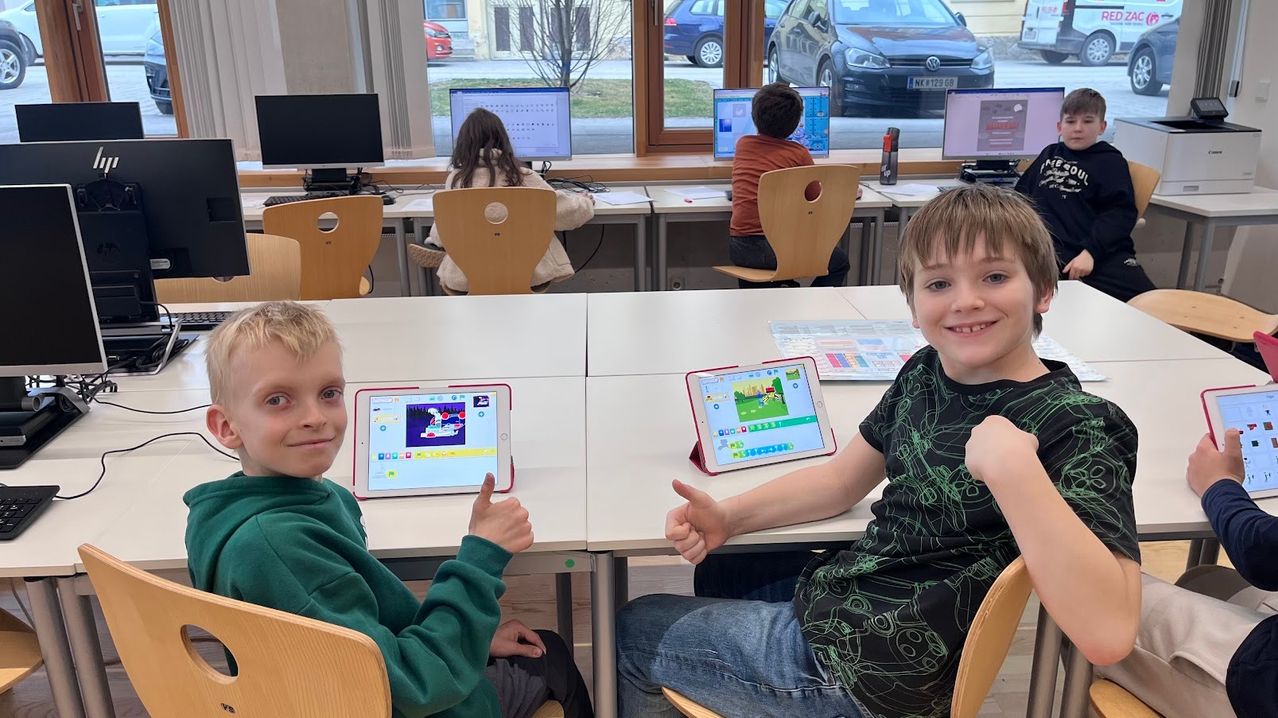 Zwei Jungen benutzen Tablets in einem Klassenzimmer. Andere Schüler sitzen an Schreibtischen mit Computern. Draußen sind zwei Autos geparkt.