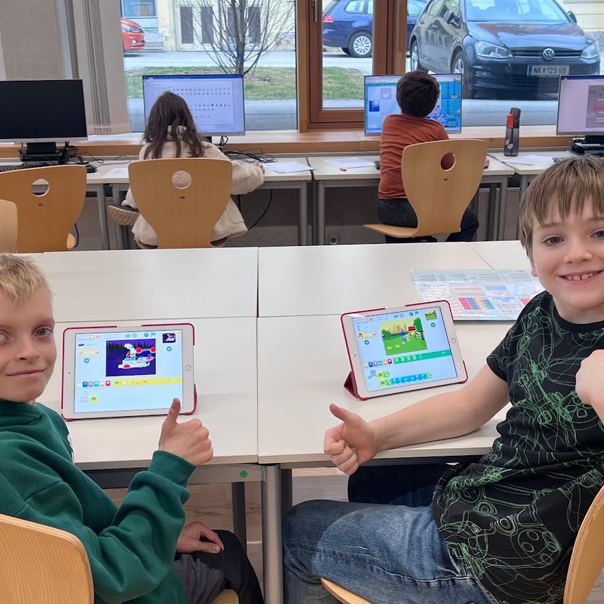 Zwei Jungen benutzen Tablets in einem Klassenzimmer. Andere Schüler sitzen an Schreibtischen mit Computern. Draußen sind zwei Autos geparkt.
