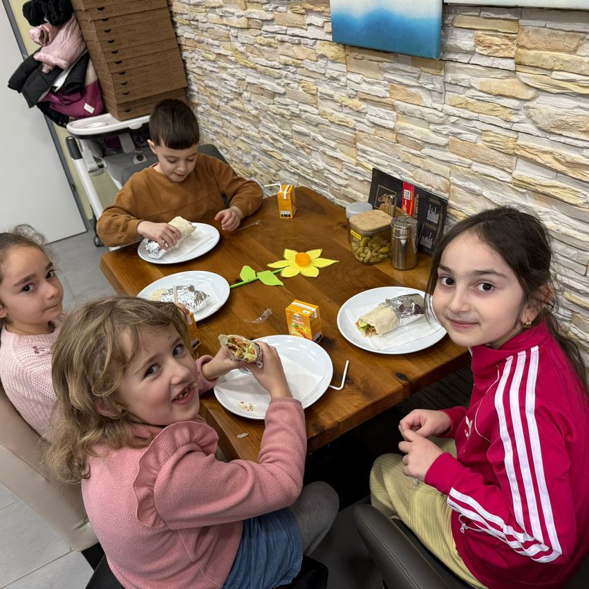 Vier Kinder sitzen um einen Holztisch herum. Sie genießen Essen und lächeln. Hinter ihnen befindet sich eine Ziegelwand mit dekorativen Gegenständen.