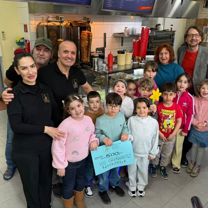 Eine Gruppe von Menschen, Erwachsene und Kinder, posiert für ein Foto in einer Bäckerei. Die Erwachsenen lächeln, und die Kinder halten ein blaues Schild mit weißen Buchstaben.