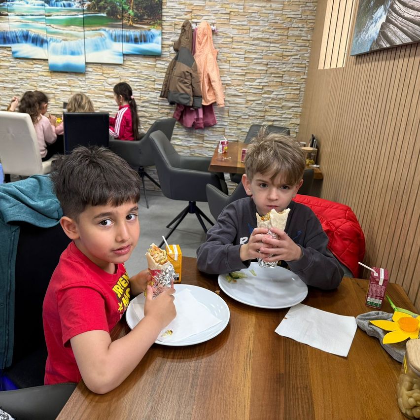 Zwei Jungen essen Burritos in einem Restaurant. Einer trägt ein rotes T-Shirt und der andere ein graues Sweatshirt. Sie sitzen an einem Tisch mit Tellern, Servietten und Getränken. Im Hintergrund sitzen andere Leute an Tischen und Jacken hängen an der Wand.