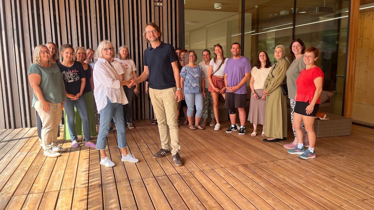 Eine Gruppe von Menschen steht auf einer Holzterrasse, lächelt und posiert für ein Foto. Ein Mann und eine Frau schütteln sich die Hände in der Mitte. Hinter ihnen stehen andere und schauen zu. Die Umgebung ist hell, mit einem modernen Gebäude im Hintergrund.