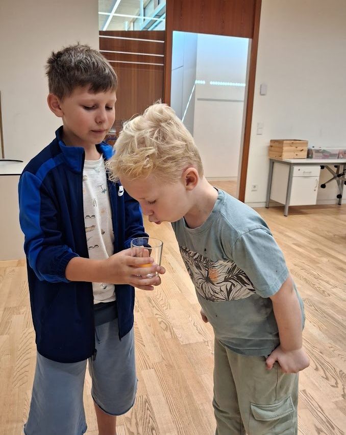 Two young boys are standing in a room, one holding a glass of liquid while the other boy is smelling it.