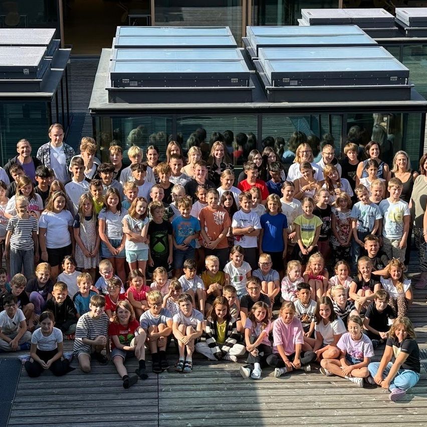 Eine große Gruppe von Kindern und Erwachsenen versammelt sich für ein Foto vor einem modernen Gebäude mit Glaswänden und Solarpanelen auf dem Dach.
