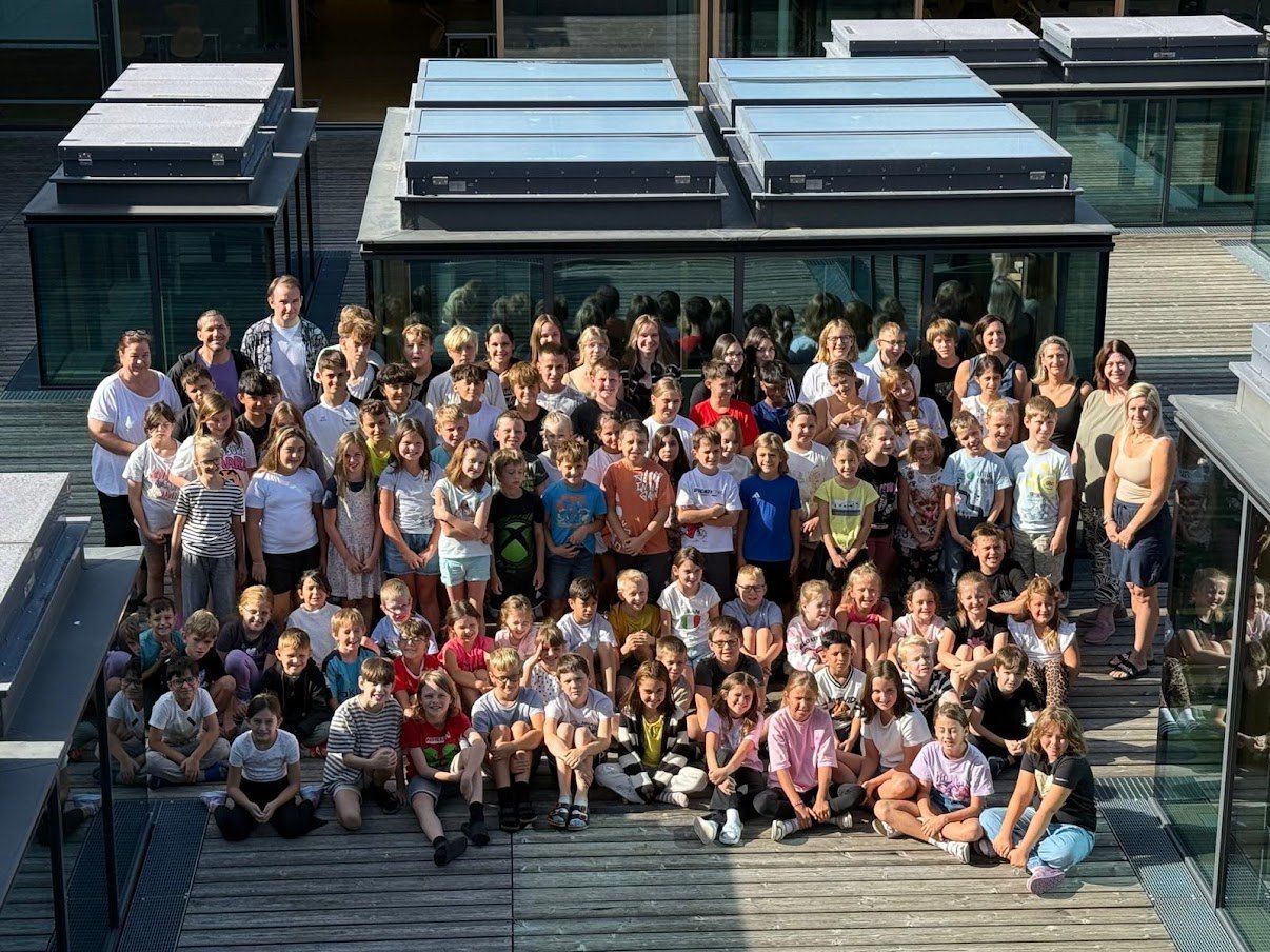 A large group of children and adults are gathered for a photo outside a modern building with glass walls and solar panels on the roof.