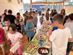 Eine Gruppe von Kindern und Erwachsenen versammelt sich um einen langen Tisch voller verschiedener Speisen. Einige essen, während andere stehen und reden. Der Raum hat große Fenster und einen Holzboden.