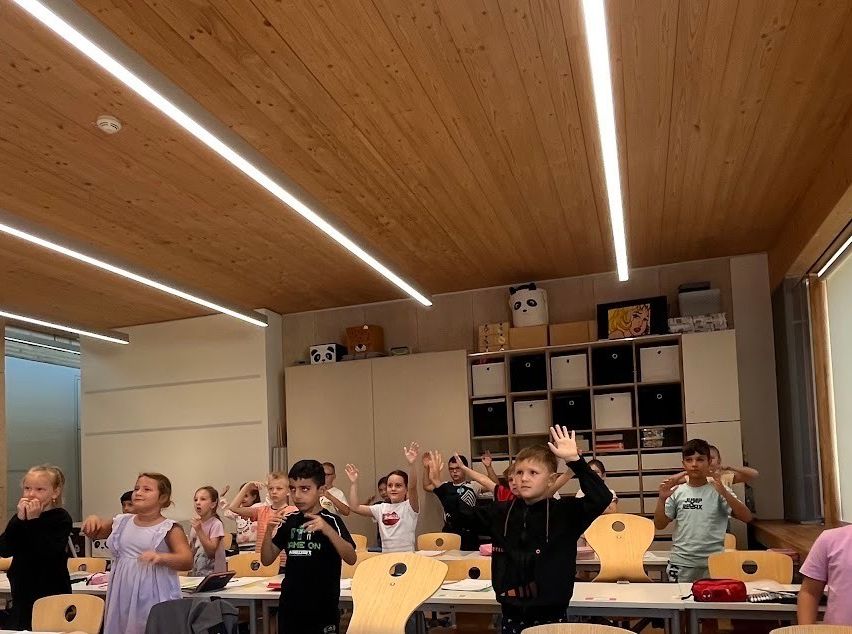 A group of children in a classroom, some standing and raising their hands, others seated at desks. The room has wooden ceilings and walls, with various items on a shelf in the background.