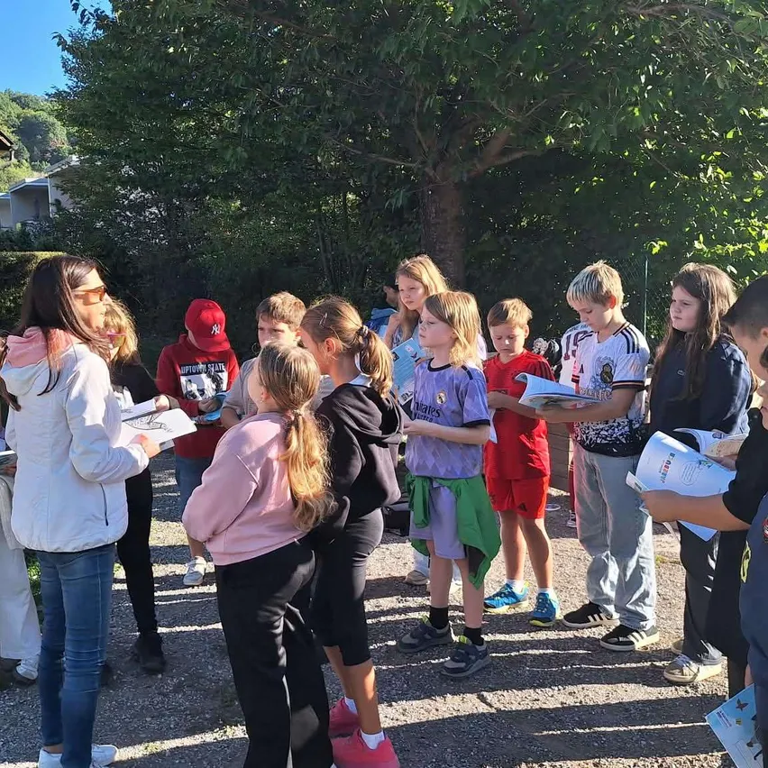 Eine Gruppe von Kindern und Erwachsenen versammelt sich im Freien, wahrscheinlich für eine Schulaktivität, wobei einige Bücher halten und andere zuschauen.
