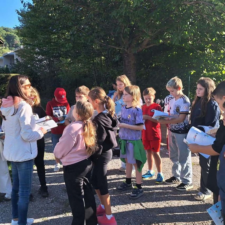 Eine Gruppe von Kindern und Erwachsenen versammelt sich im Freien, wahrscheinlich für eine Schulaktivität, wobei einige Bücher halten und andere zuschauen.