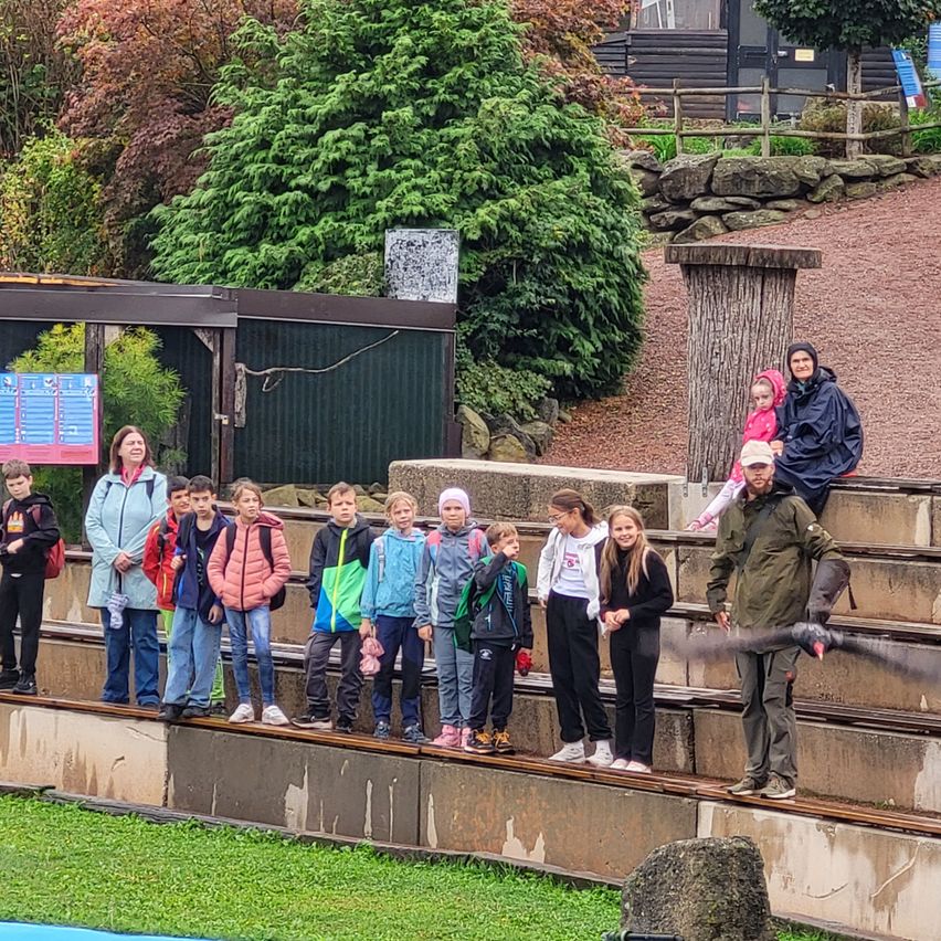 Eine Gruppe von Kindern und Erwachsenen steht auf Betontribünen, einige tragen Jacken und Mützen. Eine Frau hält einen Vogel und ein Kind sitzt auf den Schultern eines Mannes.
