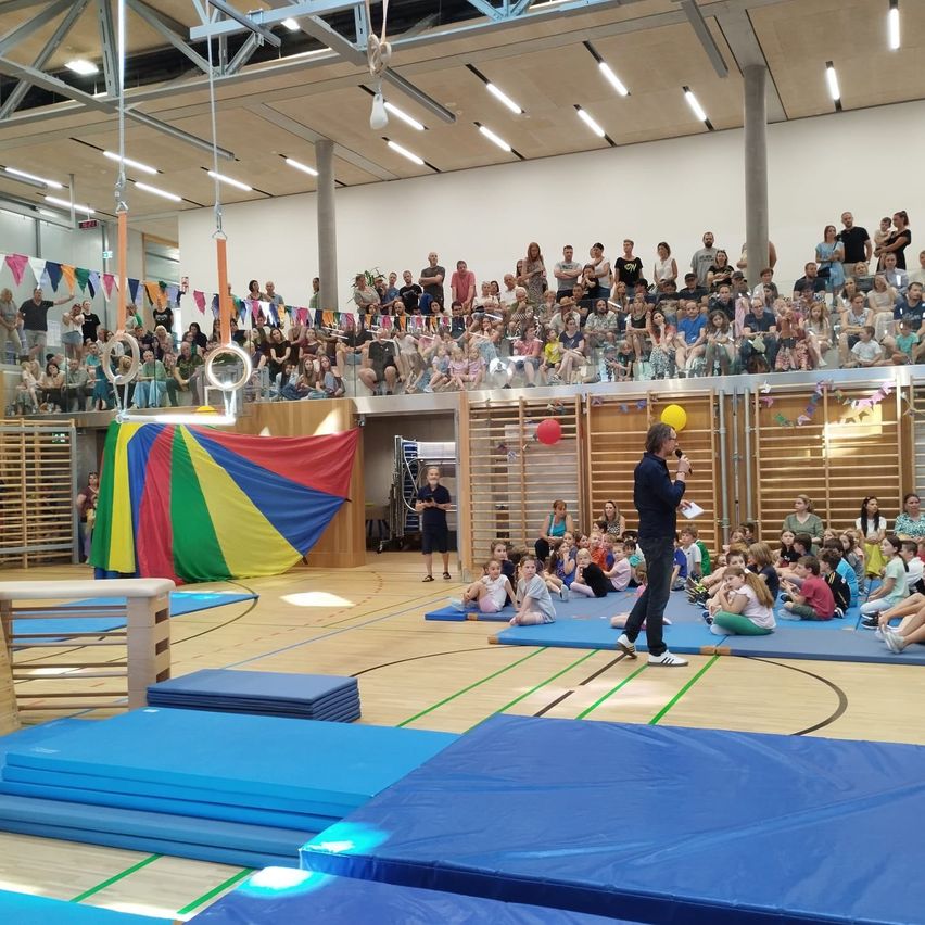Eine Gruppe von Menschen in einer Turnhalle, ein Mann spricht zu Kindern, die auf blauen Matten sitzen, ein farbenfrohes Zelt ist in der Nähe und Erwachsene schauen von den Tribünen zu.