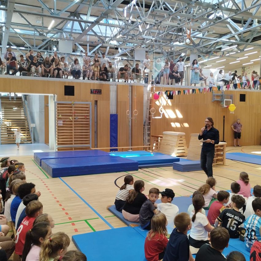Ein Mann spricht zu einer Gruppe von Kindern, die in einer Turnhalle mit blauen Matten und Holzwänden sitzen. Zuschauer sind auf der Galerie oben.